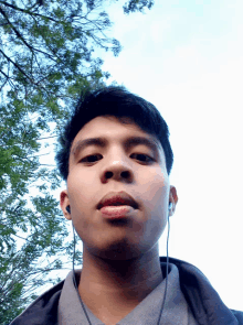 a young man wearing ear buds looks at the camera with trees in the background