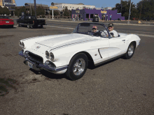 two men are sitting in a white sports car in front of a purple building that says popcorn