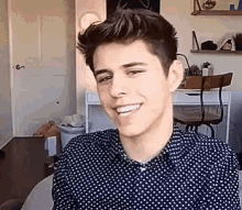 a young man in a polka dot shirt is smiling while sitting in a chair in front of a desk .