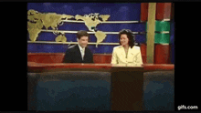 a man and a woman are sitting at a news desk with a map of the world behind them .