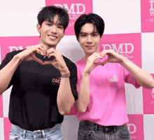 two men are making a heart shape with their hands in front of a pink wall with the word dmd on it .