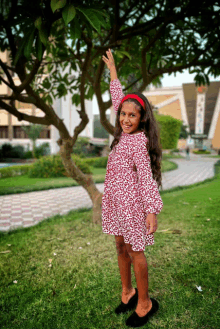 a little girl in a red dress stands in front of a tree with her hand up