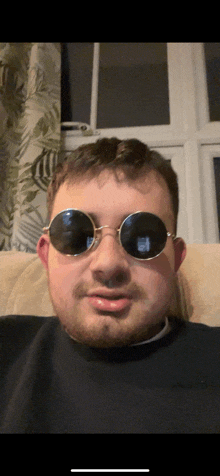 a man wearing round sunglasses sits on a couch in front of a window