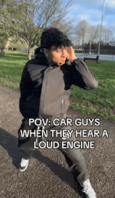 a man wearing a black jacket covering his ears with his hands while standing in a park