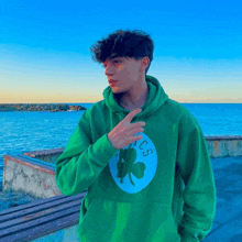 a young man wearing a green hoodie with a clover on it stands in front of the ocean