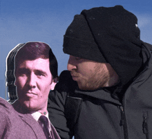 a man wearing a black beanie stands next to a cut out of a man 's face