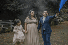 a man in a suit holds a blue smoke bomb while standing next to a pregnant woman and a little girl