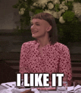 a woman with a shaved head is sitting at a table with the words " i like it " written above her