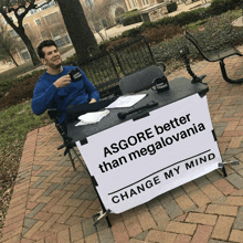 a man sits at a table with a sign that says asgore better than megalomania change my mind