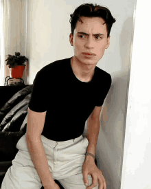 a young man wearing a black shirt and white pants is leaning against a white wall