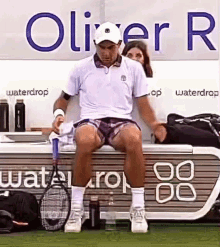 a man is sitting on a bench with a tennis racquet in front of a sign that says waterdrop .