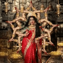 a woman in a red saree is standing in front of a statue of a goddess with many arms .