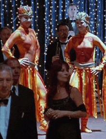 a woman in a black dress stands in front of a group of dancers in orange costumes