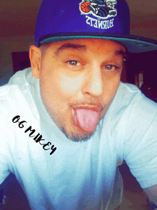 a man wearing a purple hat and a white shirt with og mikey written on it sticking out his tongue