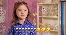 a little girl in a blue shirt is making a funny face in front of a shelf full of crowns .