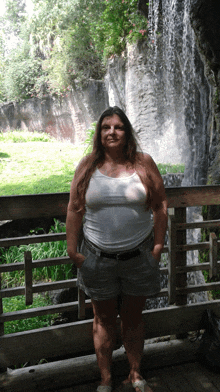 a woman stands in front of a waterfall wearing shorts and a tank top