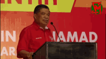 a man in a red shirt is giving a speech at a podium in front of a sign that says in jamad