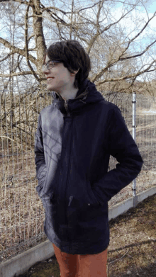 a young man wearing glasses and a black jacket stands next to a fence