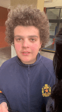 a young man with curly hair wears a blue jacket with a star on the front