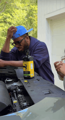 a man in a blue hat looks under the hood of a car while a bottle of black swan sits on the hood