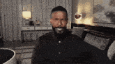 a man with a beard is sitting on a couch in a living room and smiling .