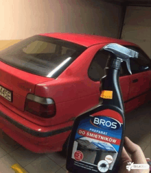 a person is holding a bottle of bros preparat do smietnikow in front of a red car