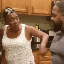 a man and a woman are standing in a kitchen talking to each other . the woman is wearing a white tank top .