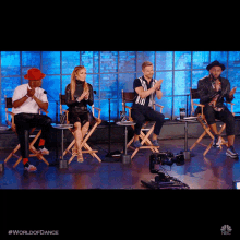 a group of people sitting in director 's chairs on a stage with the word world of dance on the bottom right