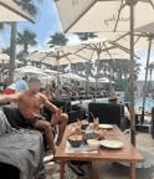 a shirtless man is sitting at a table under umbrellas at a beach restaurant .