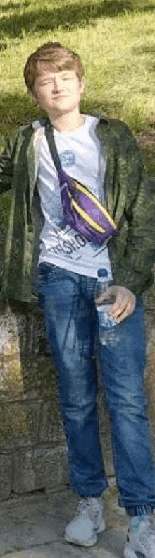 a young man is standing next to a wall holding a bottle of water and a purple fanny pack .