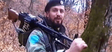 a soldier with a beard is holding a gun in the woods .