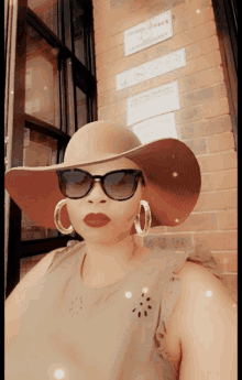 a woman wearing a hat and sunglasses stands in front of a brick wall with a sign that says " no smoking "