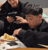 a boy is sitting at a table with a plate of food and a cell phone in his hand