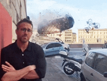 a man with his arms crossed stands in front of a car that has exploded in the background