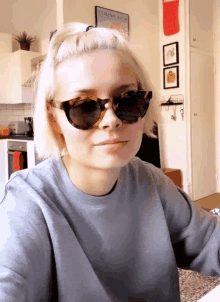 a woman wearing sunglasses looks at the camera with a poster on the wall behind her that says tourism agency