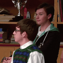 two young men are standing next to each other in front of a bookshelf with a trophy in the background