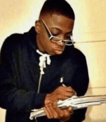 a man wearing glasses is writing in a notebook while holding a stack of money .