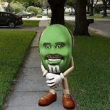 a green m & m with a beard and mustache is standing on a sidewalk next to a tree .