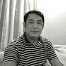 a man in a striped shirt is sitting on a couch and looking at the camera in a black and white photo .