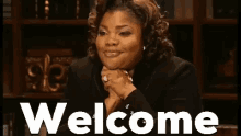 a woman is sitting at a table with her hands folded in front of a sign that says `` welcome '' .