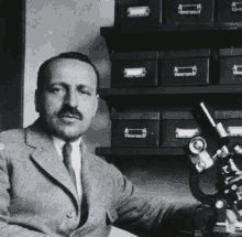 a man in a suit and tie sits in front of a microscope in a room