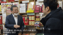 a man wearing a face mask is talking to another man in front of a shelf with chinese writing on it