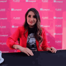 a woman in a red jacket is sitting at a table in front of a pink wall that says liverpool