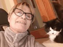 a woman wearing glasses stands next to a black and white cat looking at the camera