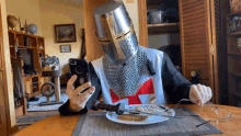 a man wearing a knight 's helmet is sitting at a table with a plate of food and a cell phone