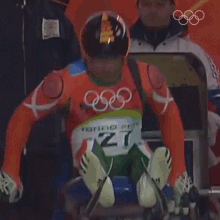 a man wearing a helmet with an arrow on it is sitting on a sled with the olympic rings in the background