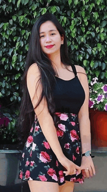 a woman in a black top and floral skirt stands in front of flowers