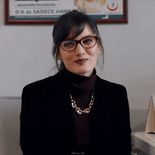 a woman wearing glasses and a necklace stands in front of a framed sign that says 0 6 ay sadece anne