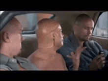 three bald men are sitting in the back seat of a car looking at a cell phone .