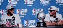 two basketball players are sitting at a table with a trophy in front of a wall that says eastern finals